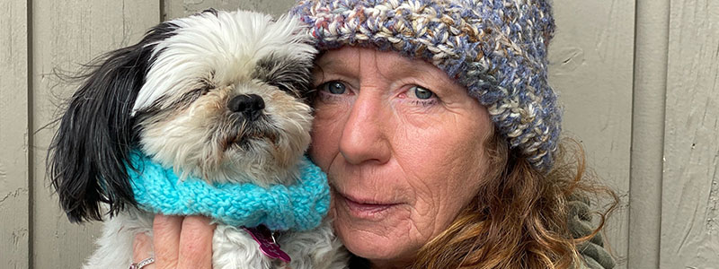 Woman wearing a knit hat holding a small dog close to her face, standing outdoors against a wooden wall.